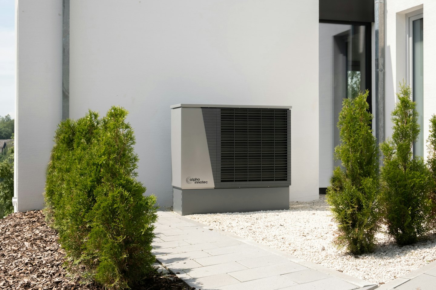 Außeneinheit einer Wärmepumpe neben einem weißen Gebäude an einem sonnigen Tag.